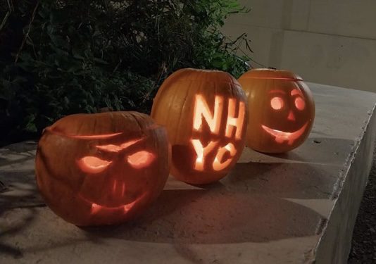 Three orange pumpkins with carvings and candles glowing from the inside, resting on a wall. Closest pumpkin has a scary face with fangs, middle pumpkin has NHYC letters, and furthest pumpkin has a happy face.